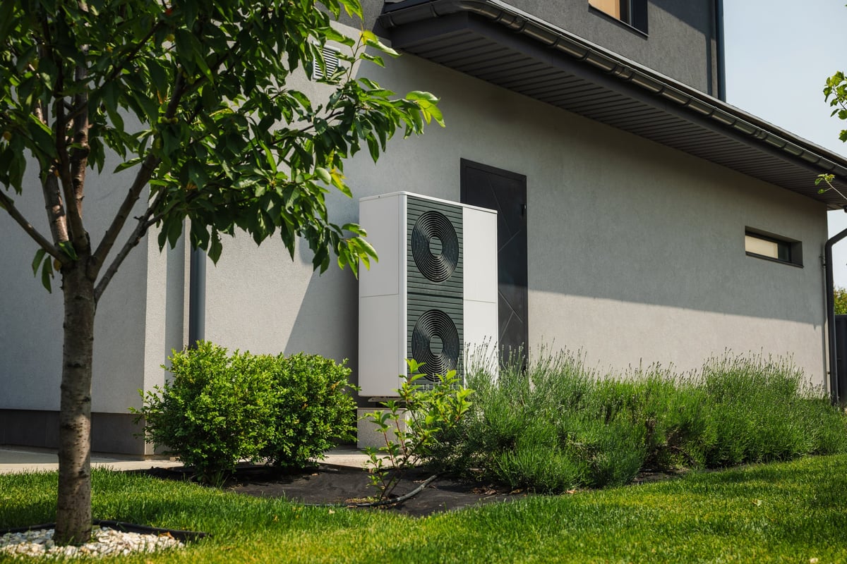 Modern heat pump unit installed next to a residential house wall, surrounded by green lawn, bushes, and young trees in a landscaped yard
